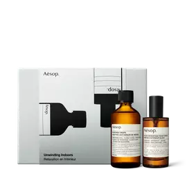 Deux flacons Aesop ambres avec bouchons noirs se tiennent devant une boite argentee ornee d’illustrations de flacons noirs.