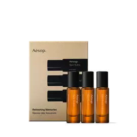 Trois flacons de parfum Aesop ambres, coiffes de hauts bouchons noirs, se tiennent devant un etui dore orne d’illustrations de flacons.