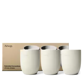 Trois contenants de bougies Aesop couleur creme sont alignes devant une boite doree ornee d’images de bougies assorties.