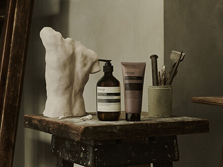 Aesop Antithesis Intense Body Cleanser pump bottle and tube beside a white torso sculpture and brushes on a wooden work table.