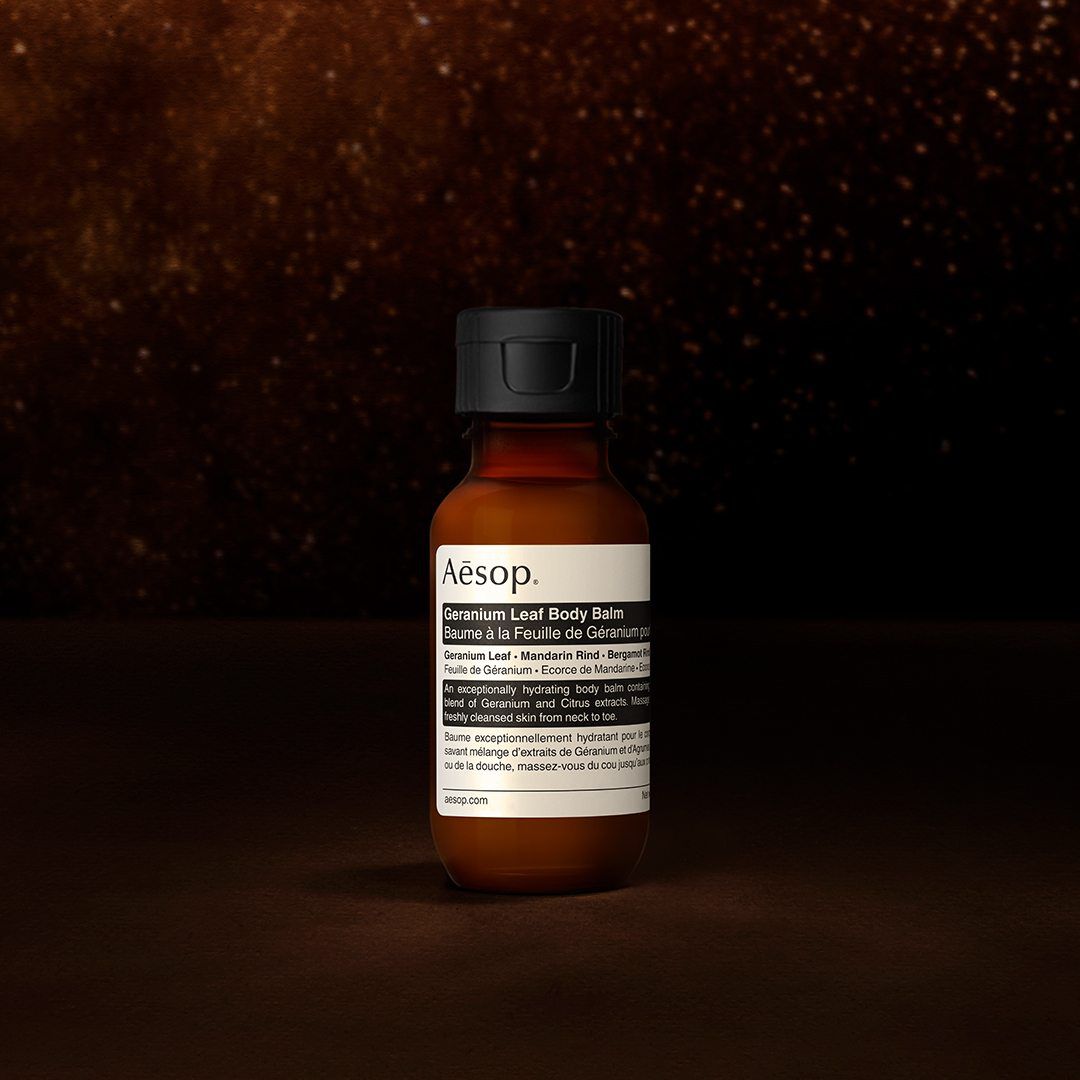 Eine Aesop Geranium Leaf Korperbalsam-Flasche vor einem dunklen, strukturierten Hintergrund.