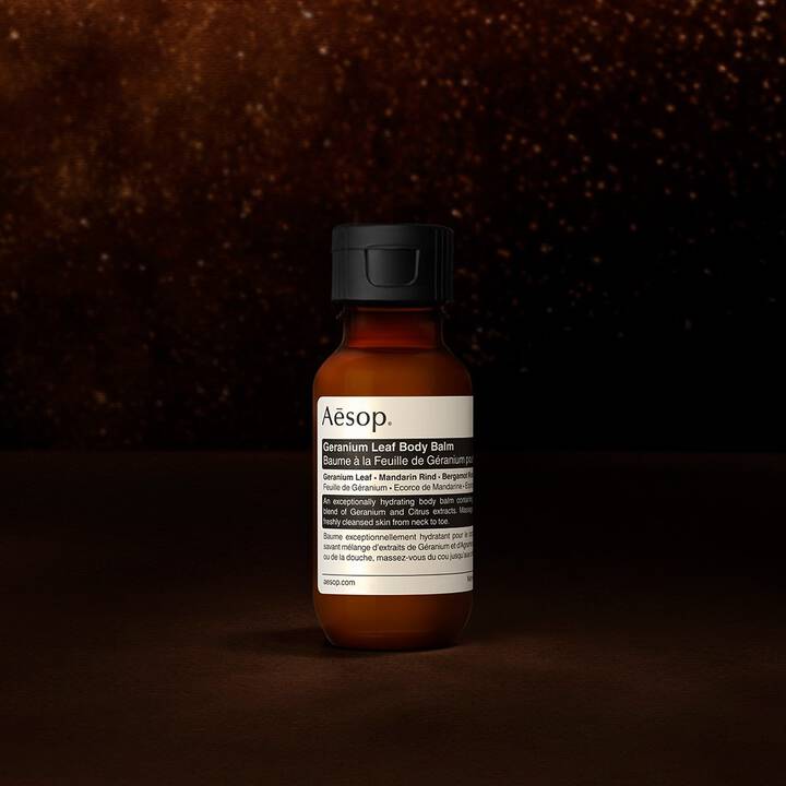 Eine Aesop Geranium Leaf Korperbalsam-Flasche vor einem dunklen, strukturierten Hintergrund.