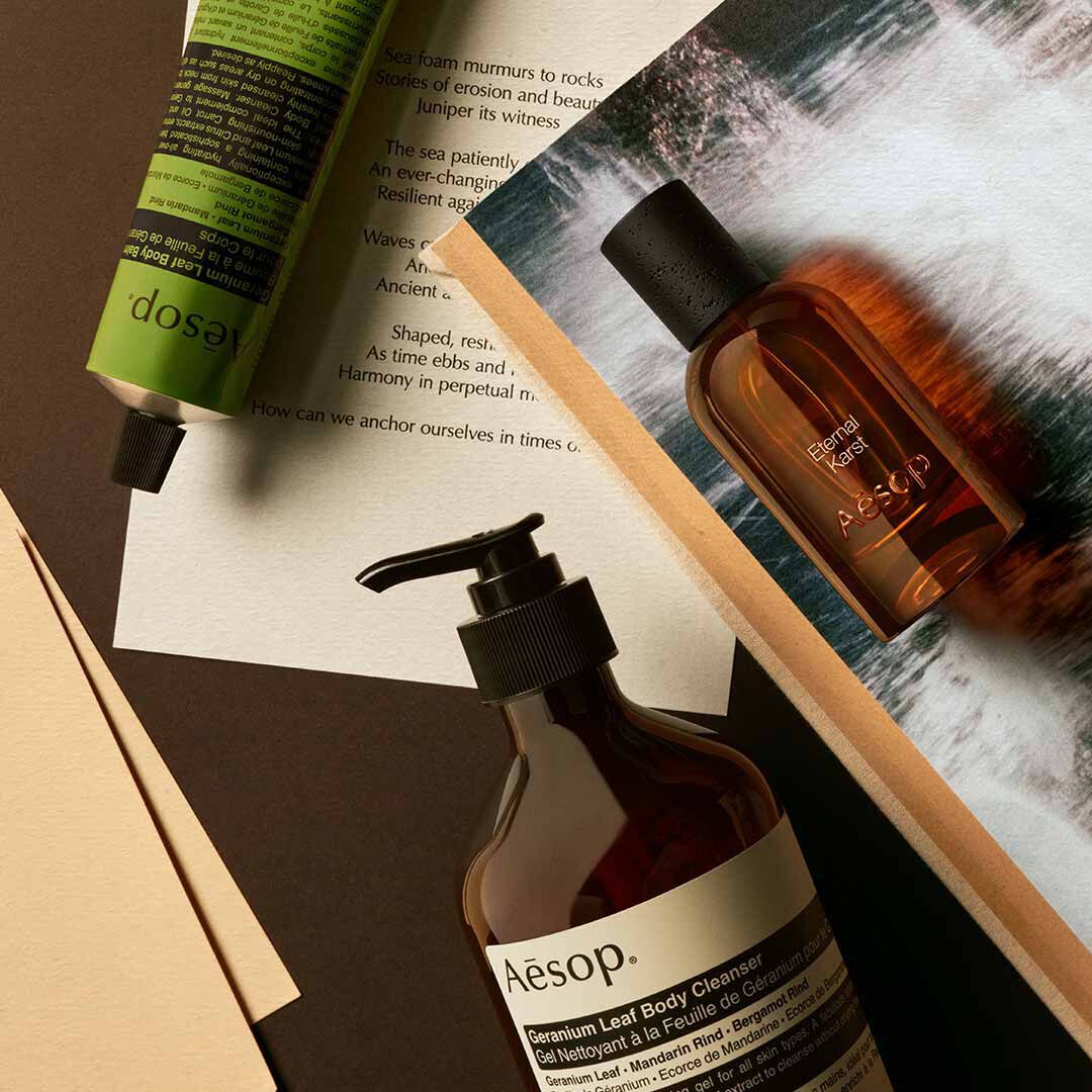 Aesop Eternal Karst Flakon mit Pumpspender und grunem Schlauch, arrangiert auf Papieren mit Bildern von schaumendem Wasser.