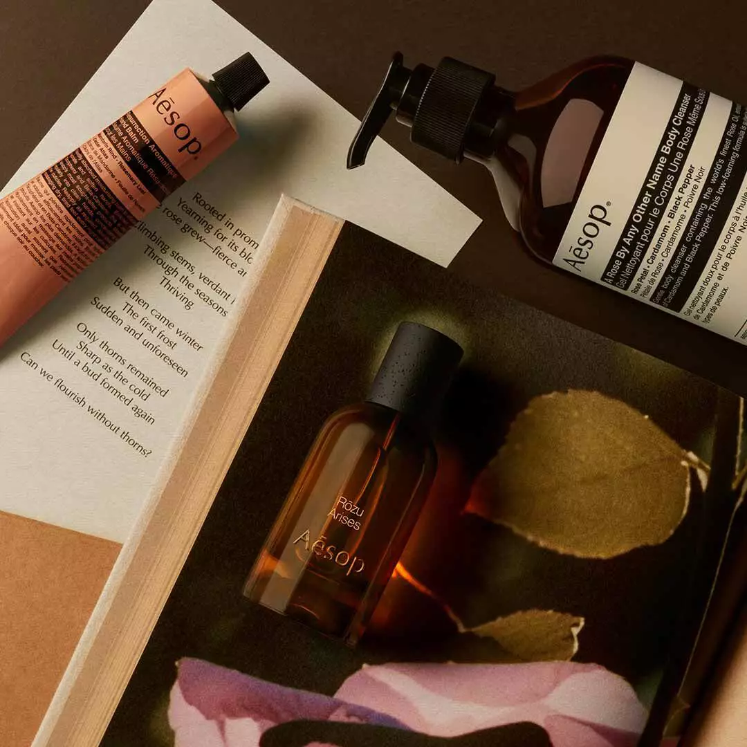 Flacon Aesop Rozu Arises avec flacon-pompe et tube couleur peche, disposes sur des papiers avec des petales roses et une feuille.