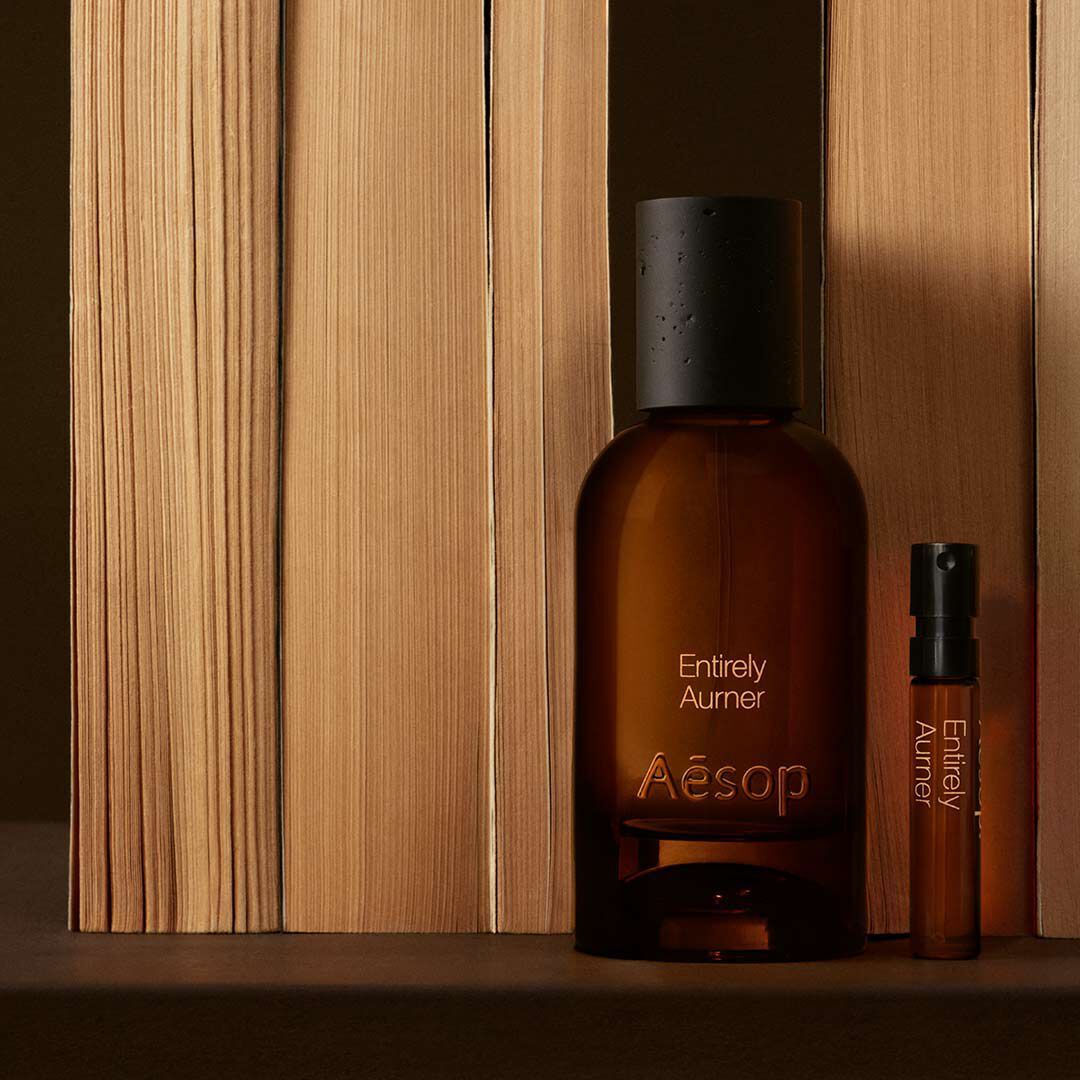 Eine Bernsteinglasflasche des Parfums Aesop Entirely Aurner und ein passendes Probchen stehen aufrecht neben vertikal angeordneten Buchern auf einer dunklen Oberflache.