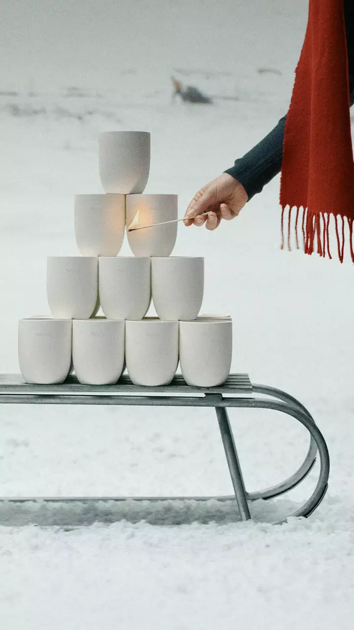 A hand holds a burning match beside white vessels stacked on a metal sledge in snow, with a red fringed scarf to the right.